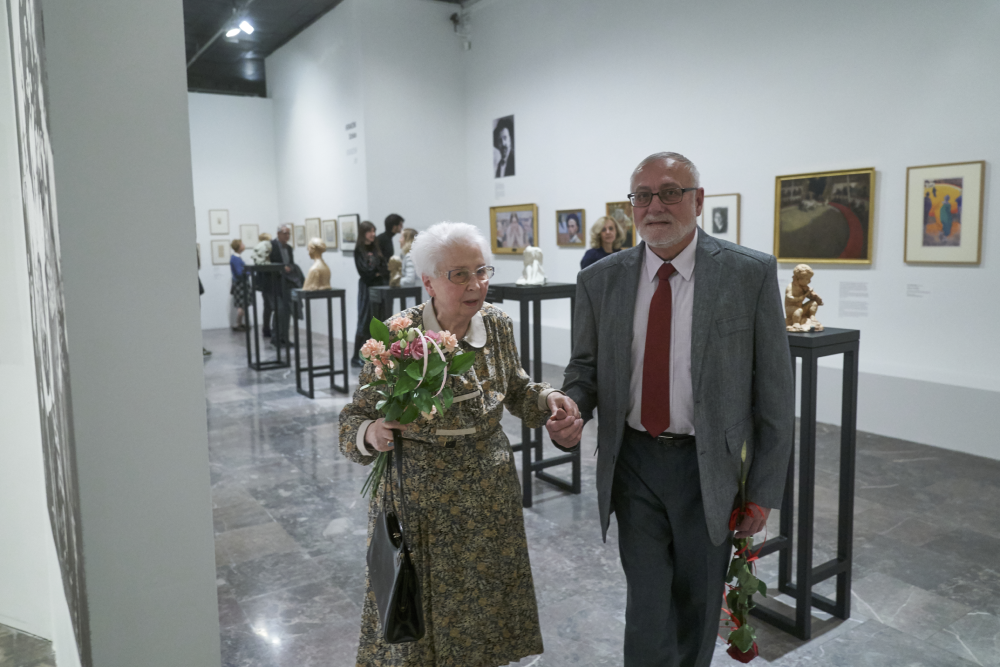 Starsza para trzymająca się za ręce przechodzi przez przestronne białe wnętrze galerii sztuki; kobieta z białymi włosami w kwiecistej sukni trzyma bukiet róż i czarną torebkę, mężczyzna w szarym garniturze i czerwonym krawacie niesie pojedyncze czerwone róże, wokół stoją czarne postumenty z popiersiami, obrazy wiszą na białych ścianach, a na błyszczącej marmurowej posadzce widać innych zwiedzających.