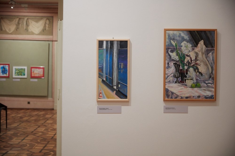 Widok wnętrza galerii: na białej ścianie przy otwartym przejściu wiszą dwie drewniane ramy — wąski pionowy obraz o dominujących odcieniach niebieskiego z widokiem okna i małym pomarańczowym pachołkiem u dołu oraz większy obraz martwej natury z wazonami, kwiatami i dwoma zielonymi owocami na zasłonie, a po lewej widać przejście do sąsiedniej sali z trzema kolorowymi obrazami na zielonej ścianie, drewnianą podłogą i dekoracyjnym gzymsem.