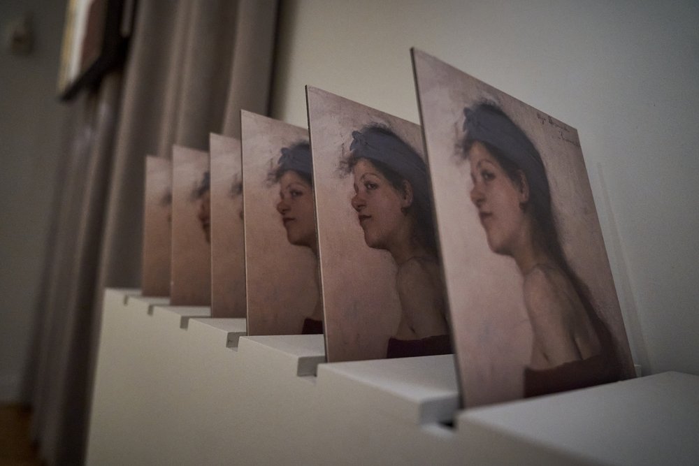 Kilka identycznych wydruków przedstawiających portret kobiety w profilu z opaską na głowie ustawionych jeden za drugim na białej półce w diagonalnej perspektywie, z rozmytym tłem i zasłoną po lewej.