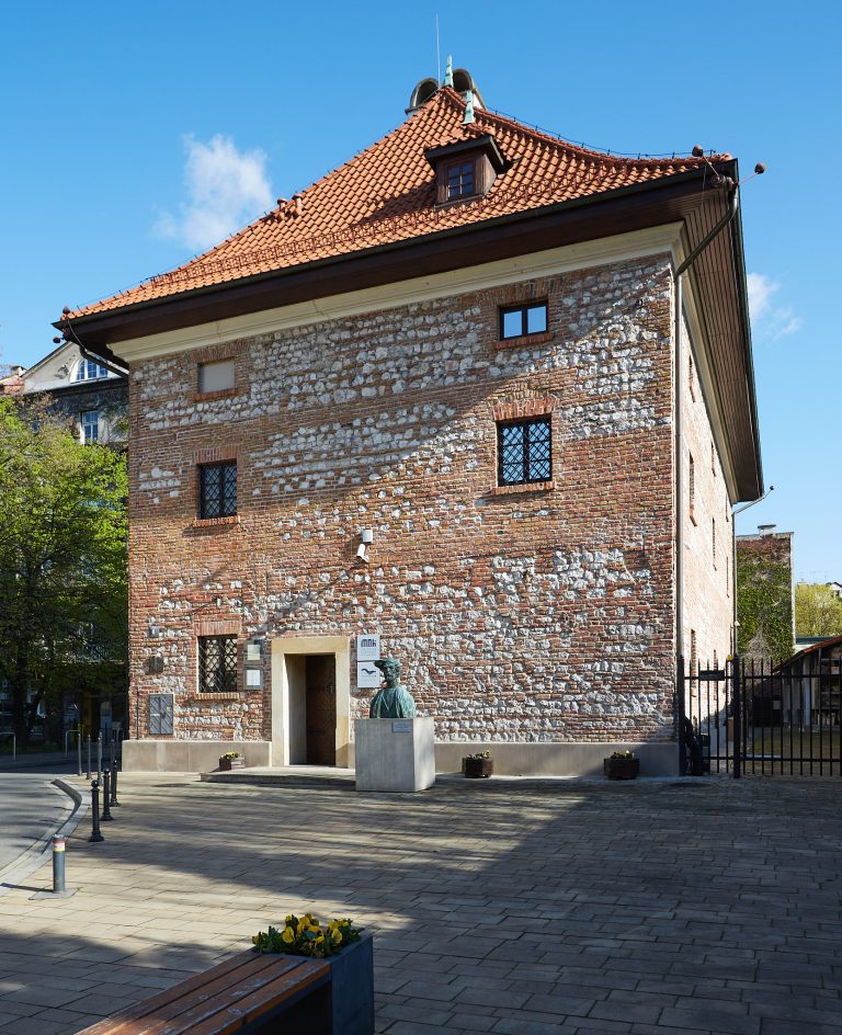 Kamienno-ceglana elewacja budynku z czerwonym dachem krytym dachówką i niewielkim oknem dachowym, metalowe kraty w oknach, przed wejściem postument z popiersiem, brukowany plac z ławką i donicami z kwiatami oraz ogrodzeniowa brama po prawej, na tle drzew i błękitnego nieba.