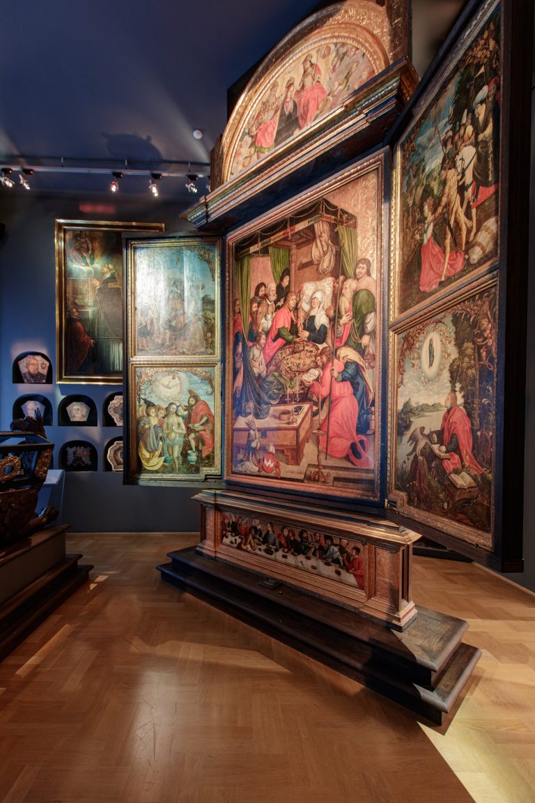 Wnętrze muzeum z dużym, otwartym drewnianym ołtarzem-tryptykiem o złoconych ramach i wielobarwnych malowanych panelach ukazujących liczne sceny z postaciami, ustawionym na zdobionej podstawie i stopniach na parkietowej podłodze, z bogato dekorowanymi łukami i bocznymi skrzydłami, oświetlonym sufitowymi reflektorami; po lewej widoczne mniejsze oprawione obrazy i ceramiczne maski.