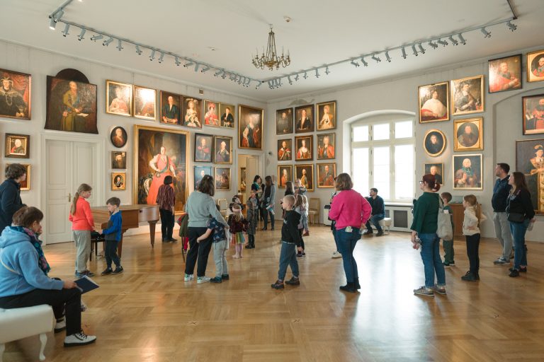 Wnętrze galerii z wysokim sufitem i żyrandolem, ściany wypełnione licznymi portretami w ramach podświetlonymi reflektorami, duże okno, drewniana parkietowa podłoga oraz grupa zwiedzających — dorośli i dzieci — stojących i poruszających się po sali przy fortepianie i ławkach.