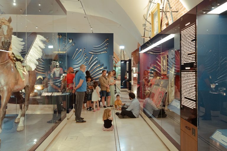 Wnętrze muzeum z długim, wąskim korytarzem przeszklonych gablot po obu stronach; po lewej częściowo widoczny model konia ozdobiony białym pióropuszem, na tylnej ścianie ułożone wachlarzowo szable i topory, w gablotach z prawej tkaniny, zbroja i portret w złotej ramie oraz stojąca pionowa tablica z tekstem, a na środku grupa dorosłych i dzieci oglądająca eksponaty i siedząca na podłodze, z refleksami świetlnymi na szybach.