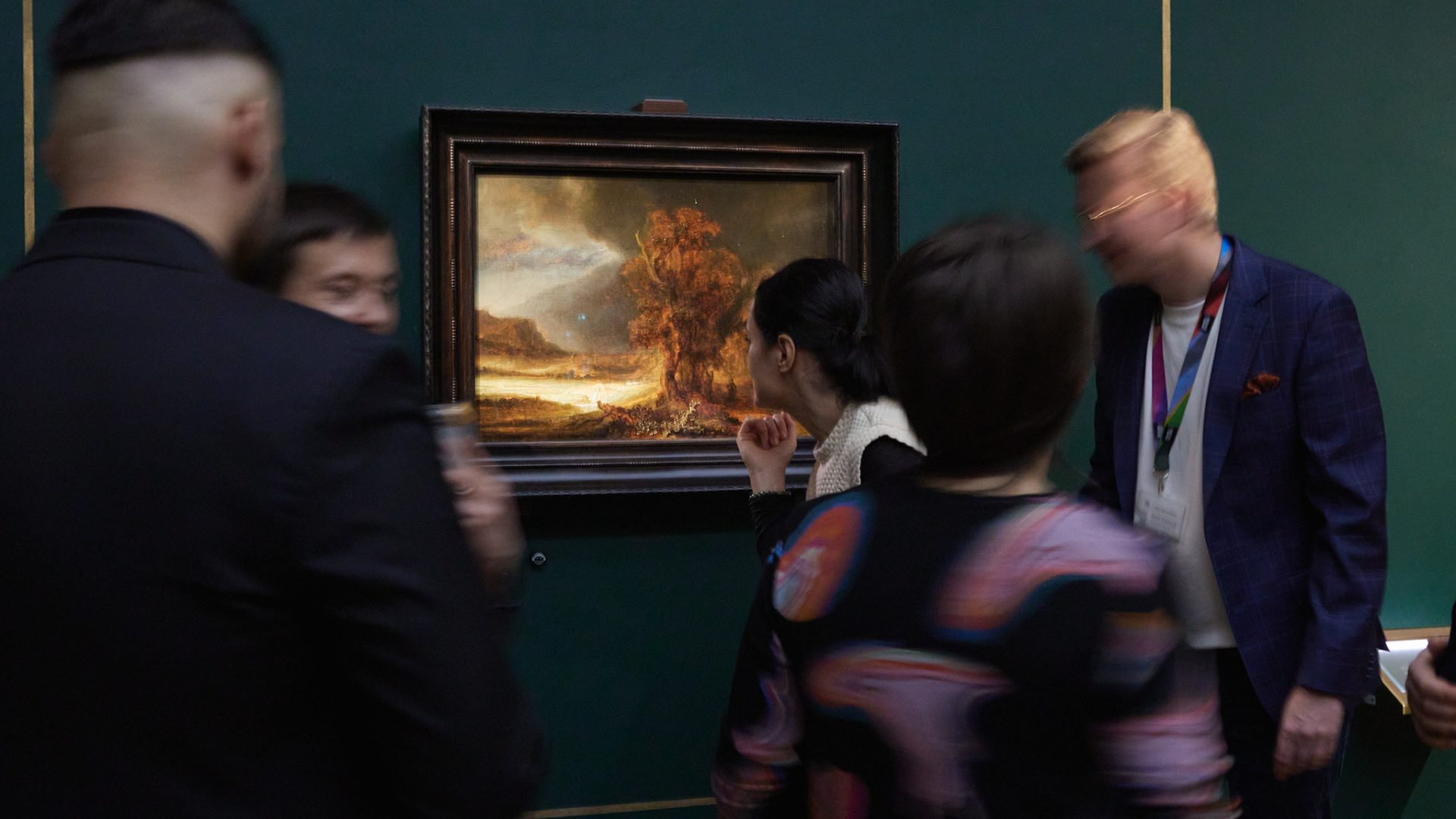 Kilka rozmytych osób oglądających mały, oprawiony pejzaż z dużym drzewem i jasnym niebem zawieszony na ciemnozielonej ścianie z cienkimi złotymi listwami; kobieta pochylona blisko obrazu pośrodku i mężczyzna w niebieskiej marynarce z identyfikatorem po prawej.