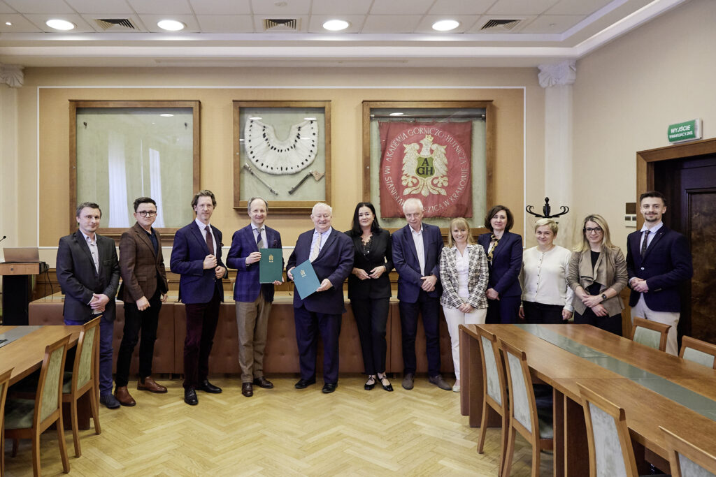 Trzynastoosobowa grupa osób w formalnym ubraniu stoi w jednym rzędzie w sali konferencyjnej przed trzema oprawionymi eksponatami na ścianie — pośrodku półokrągły metalowy element i narzędzia, z prawej czerwony sztandar z herbem i literami "AGH" — dwoje mężczyzn trzyma zielone teczki, wokół drewniane stoły i krzesła, parkiet, sufitowe oświetlenie i po prawej drzwi z zielonym znakiem "WYJŚCIE EWAKUACYJNE".