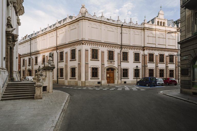 Ujęcie uliczne z narożnym, ozdobnym budynkiem o jasnej, rzeźbionej górnej elewacji i ceglanych detalach na parterze, drewnianymi drzwiami i wieloma oknami, kamiennymi schodami i balustradą ze rzeźbami po lewej, kilkoma zaparkowanymi samochodami po prawej, pustą jezdnią z oznaczeniami poziomymi oraz fragmentem nieba w tle, widoczny znak uliczny Pijarska.