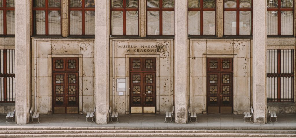 Fasada budynku z trzema drewnianymi drzwiami z ozdobnymi szybami ustawionymi w symetrycznych niszach między masywnymi kamiennymi filarami, nad środkowymi drzwiami napis "MUZEUM NARODOWE W KRAKOWIE", duże prostokątne okna z czerwonymi ramami powyżej, zabrudzona kamienna elewacja, schody i metalowe ławki przed wejściami.
