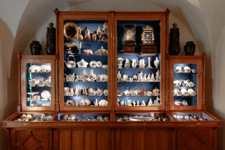 Drewniana, trzyczęściowa witryna z przeszklonymi półkami i niebieskim wnętrzem, oświetlona i wypełniona porcelaną, filiżankami, talerzami, dzbankami, srebrnymi naczyniami, muszlą, figurkami i dwoma ozdobnymi zegarami, z poziomymi przeszklonymi gablotami na dole z drobnymi przedmiotami oraz dwoma wysokimi ciemnymi wazonami na górze, na tle jasnego sklepionego sufitu.