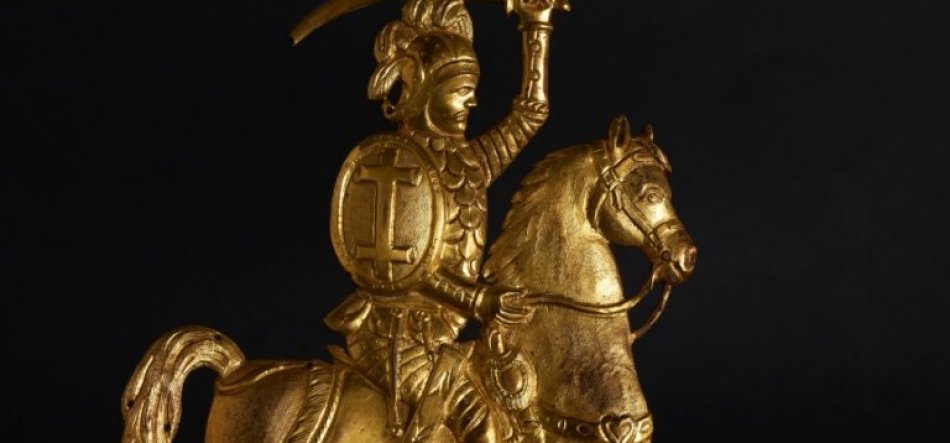 Złocona figurka zbrojnego rycerza w hełmie z pióropuszem jadącego na koniu, unoszącego miecz i trzymającego tarczę z wyraźnym krzyżem na czarnym tle.