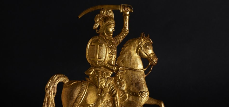Złota figurka jeźdźca na koniu na ciemnym tle — rycerz w hełmie z pióropuszem trzyma uniesiony zakrzywiony miecz, ma okrągłą tarczę z wypukłym pionowym i poziomym reliefem, a koń ma ozdobną uprząż.