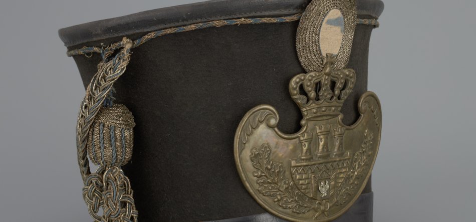 Zbliżenie czarnej filcowej czapki typu czako z dużą mosiężną plakietą przedstawiającą koronę nad trzema wieżami i liśćmi dębu oraz splątanymi złotymi i niebieskimi sznurami z chwostem po lewej, z widocznym skórzanym daszkiem u dołu.