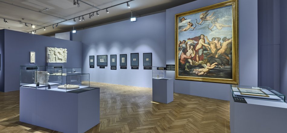 Przestronna sala wystawowa z jasnoniebieskimi ścianami i jodełkowym parkietem, sufitowymi reflektorami, dużym złoconym obrazem z grupą postaci i skrzydlatymi figurami po prawej, kilkoma przeszklonymi gablotami z eksponatami na pierwszym planie oraz reliefem i szeregiem małych oprawionych prac wiszących na lewej ścianie.