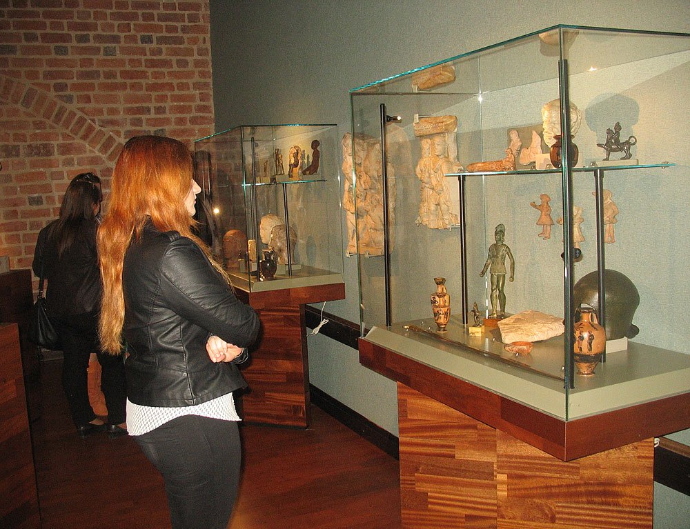 Wnętrze muzeum z dwiema zwiedzającymi: na pierwszym planie kobieta z długimi rudymi włosami i czarną skórzaną kurtką stoi bokiem przed szklanymi gablotami z antycznymi figurkami, wazami i hełmem, w tle druga osoba i ceglana ściana.