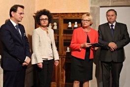 Zdjęcie czterech dorosłych stojących w rzędzie przed drewnianą witryną z ozdobami: po lewej mężczyzna w ciemnym garniturze, obok kobieta z kręconymi włosami w jasnej marynarce, dalej kobieta w czerwonej marynarce trzymająca dokumenty, a po prawej kolejny mężczyzna w ciemnym garniturze.