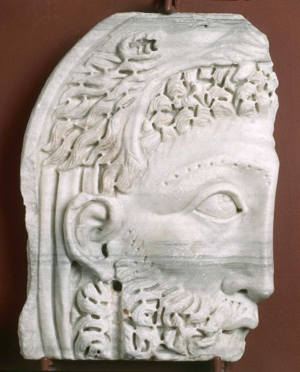 Fragment białego marmurowego reliefu ukazujący profil twarzy mężczyzny z brodą, wyraźnym uchem i okiem, z bogato zdobionymi włosami oraz ornamentami przypominającymi liście, z widocznymi otworami i pęknięciami, ustawiony na brązowym tle.