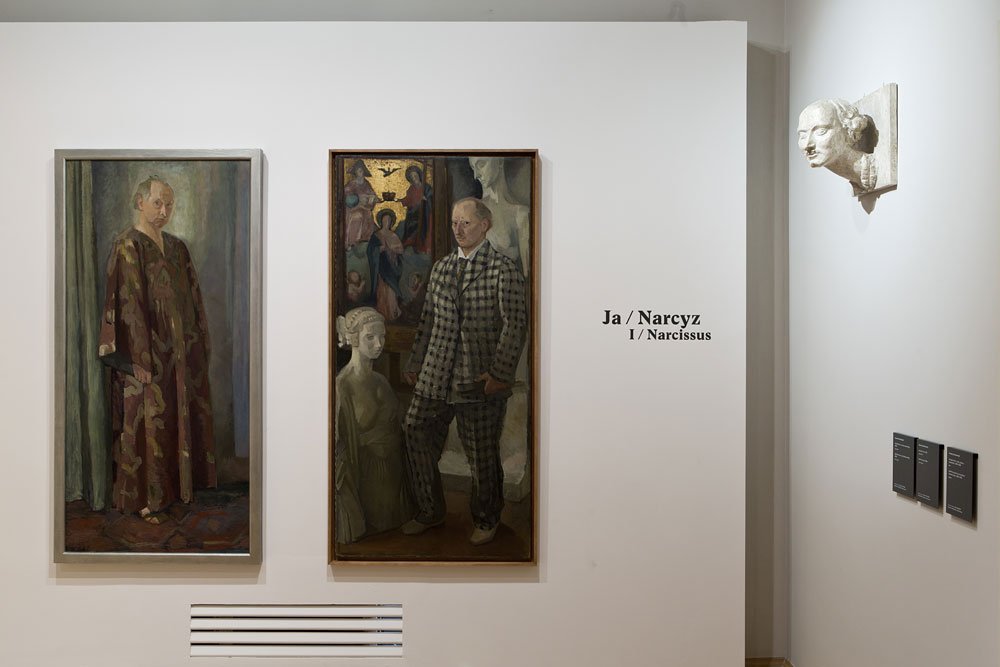 Wnętrze galerii z białą ścianą, na której wiszą dwa wysokie portretowe obrazy w ramach — po lewej postać w długiej wzorzystej szacie, pośrodku mężczyzna w kraciastej marynarce stojący obok kamiennego popiersia i ikonograficznego tła; obok na ścianie napis Ja / Narcysz I / Narcissus, po prawej przymocowana kamienna rzeźba głowy i trzy małe tabliczki informacyjne, pod obrazami kratka wentylacyjna.