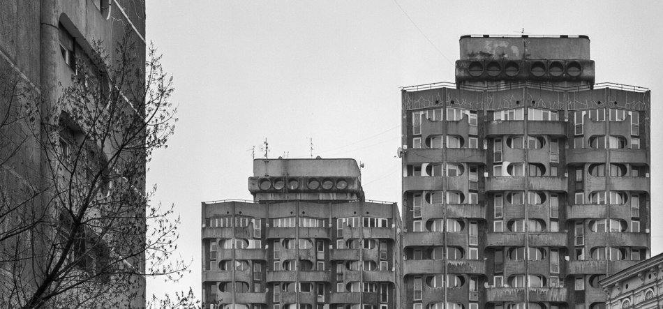 Czarno-białe zdjęcie trzech wysokich betonowych wieżowców o surowej, modułowej fasadzie z półokrągłymi wnękami balkonów, z gałęziami drzewa i fragmentem innego budynku po lewej oraz kawałkiem niższej ozdobnej fasady po prawej, na tle jasnego nieba.