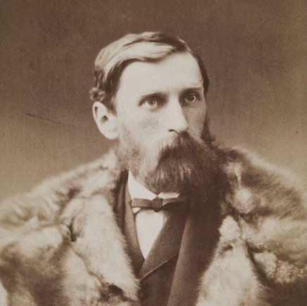 Portret w tonacji sepii w ujęciu głowa i ramiona przedstawiający mężczyznę z gęstą brodą i wąsami, ubranym w futrzane okrycie i marynarkę z muszką, patrzący nieco w prawo.