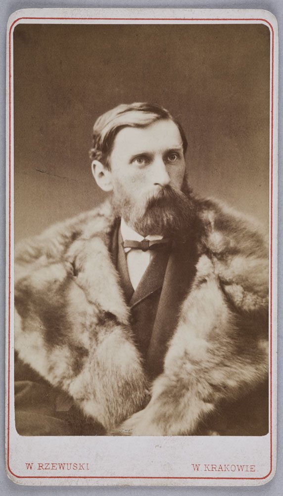 Sepia portret trzech czwartych mężczyzny z boczną przedziałką, gęstą brodą i wąsami, w ciemnym garniturze i muszce, otulonego futrzanym płaszczem, na neutralnym tle z cienką czerwoną ramką i nadrukiem u dołu W. Rzewuski W. Krakowie