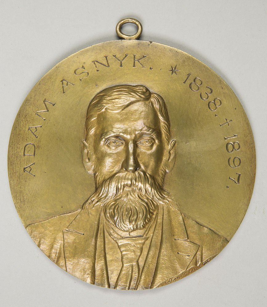 Okrągły złocisty medal z oczkowatym uchem na górze, z wypukłym portretem brodatego mężczyzny w marynarce i krawacie na środku oraz napisem na obwodzie "ADAM ASNYK" i datami "1838" i "1897".