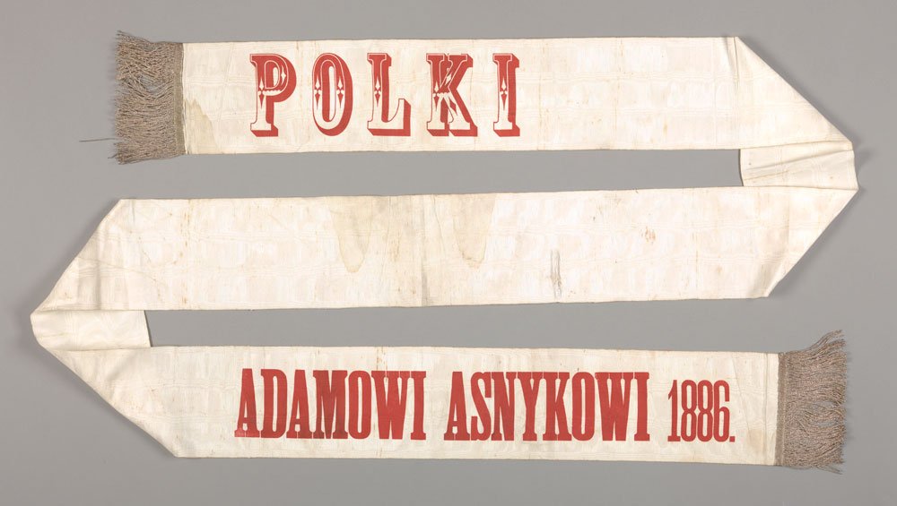 Biała szarfa rozłożona w zygzak na szarym tle, z dużymi czerwonymi literami "POLKI" na jednym końcu i napisem "ADAMOWI ASNYKOWI 1886." na drugim, obustronnie zakończona beżowymi frędzlami i z widocznymi przebarwieniami tkaniny.