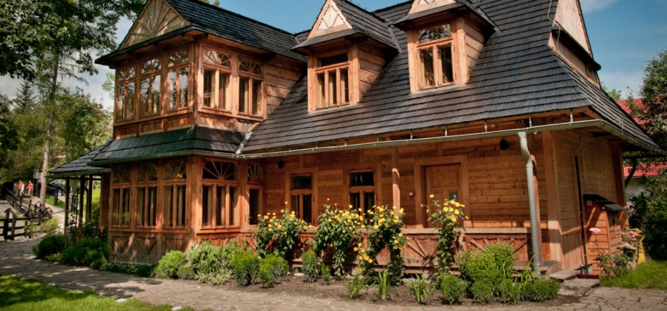 Drewniana, dwukondygnacyjna willa z ciemnym, wielospadowym dachem i dwoma lukarnami, ozdobnymi ramami okien, przeszklonym narożnym wykuszem, żółtymi kwiatami w skrzynkach przed fasadą, kamiennym chodnikiem i zadbanymi rabatami otoczona trawą i drzewami pod błękitnym niebem.