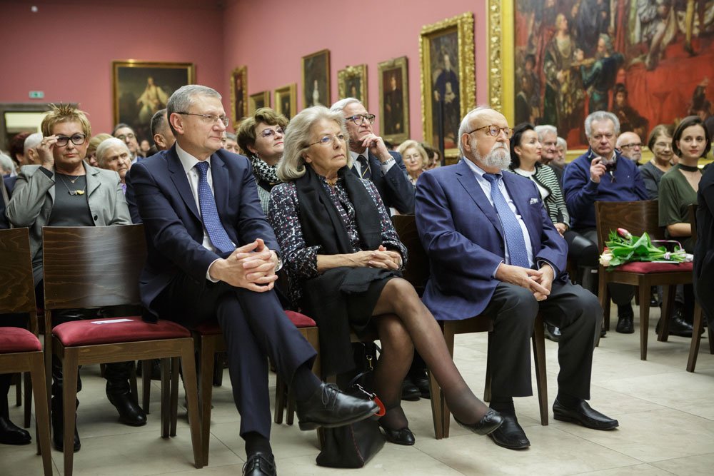 Wnętrze sali wystawowej z czerwonymi ścianami i wieloma obrazami w złotych ramach; w rzędach drewnianych krzeseł z czerwonymi poduszkami siedzi grupa ludzi, na pierwszym planie trzech starszych: mężczyzna po lewej w ciemnym garniturze i krawacie, kobieta w środku w sukience i czarnym szalu oraz mężczyzna po prawej z białą brodą, okularami i niebieską marynarką, a po prawej na krześle leży bukiet kwiatów.