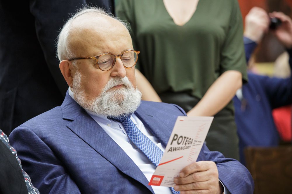 Starszy mężczyzna z białą brodą i okularami w niebieskim garniturze i krawacie siedzi i trzyma ulotkę, a w tle stoi kobieta w zielonej bluzce.