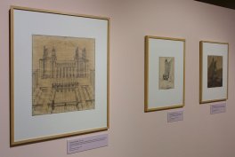 Trzy oprawione rysunki architektoniczne zawieszone w rzędzie na jasnoróżowej ścianie galerii — większy szkic budynku po lewej i dwa mniejsze po prawej, każdy w złotej ramie z podpisami pod nimi.