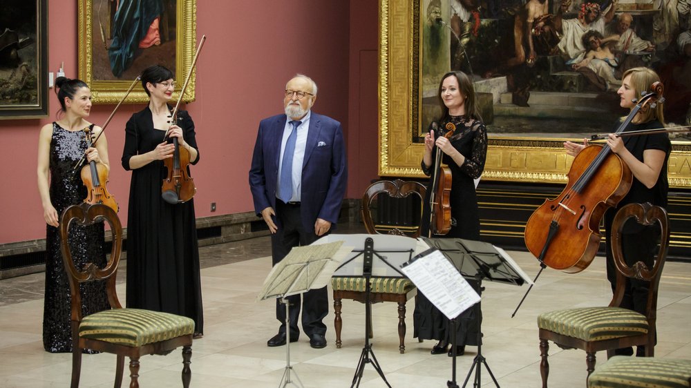 Pięć osób stoi w sali przypominającej muzeum przed dużymi złoconymi ramami na różowawych ścianach — cztery kobiety trzymają instrumenty smyczkowe (skrzypce i wiolonczelę) i starszy mężczyzna w garniturze, ustawieni w półokręgu wokół stojaków z nutami i ozdobnych krzeseł.
