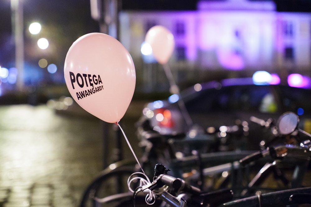 Różowy balon z czarnym napisem POTEGA AWANGARDY przywiązany do kierownicy jednego z zaparkowanych rowerów, na pierwszym planie kilka innych rowerów, w tle rozmyte światła uliczne, samochód i fioletowo oświetlony budynek.