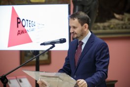 Mężczyzna w granatowym garniturze i bordowym krawacie stoi przy przezroczystym pulpicie i przemawia do mikrofonu, obok po lewej widać ekran z czerwonym trójkątem i napisem POTEGA, a w tle obraz na różowej ścianie.