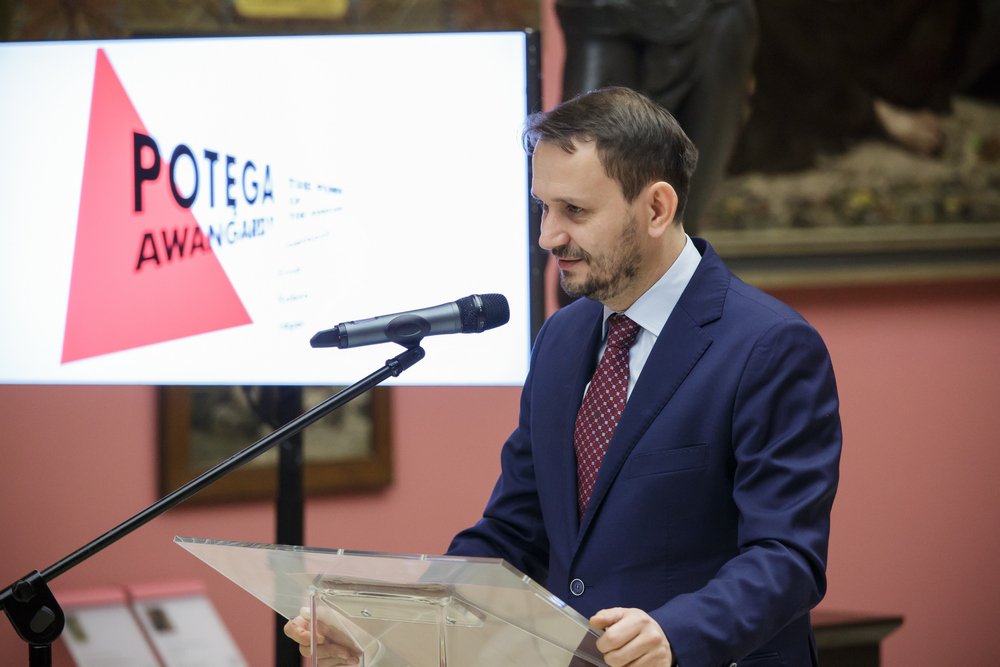 Mężczyzna w granatowym garniturze i czerwonym krawacie stoi za przezroczystą mównicą przy mikrofonie, po lewej duży ekran z czerwonym trójkątem i częścią napisu "POTEGA AWAN", a w tle różowa ściana z obrazami.