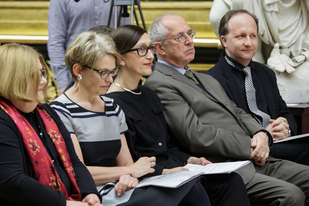 Pięć dorosłych osób siedzi w rzędzie w sali — trzy kobiety i dwóch mężczyzn w formalnym ubraniu, niektórzy w okularach, trzymają kartki z dokumentami; w tle złota dekoracja ścienna i fragment marmurowej rzeźby.