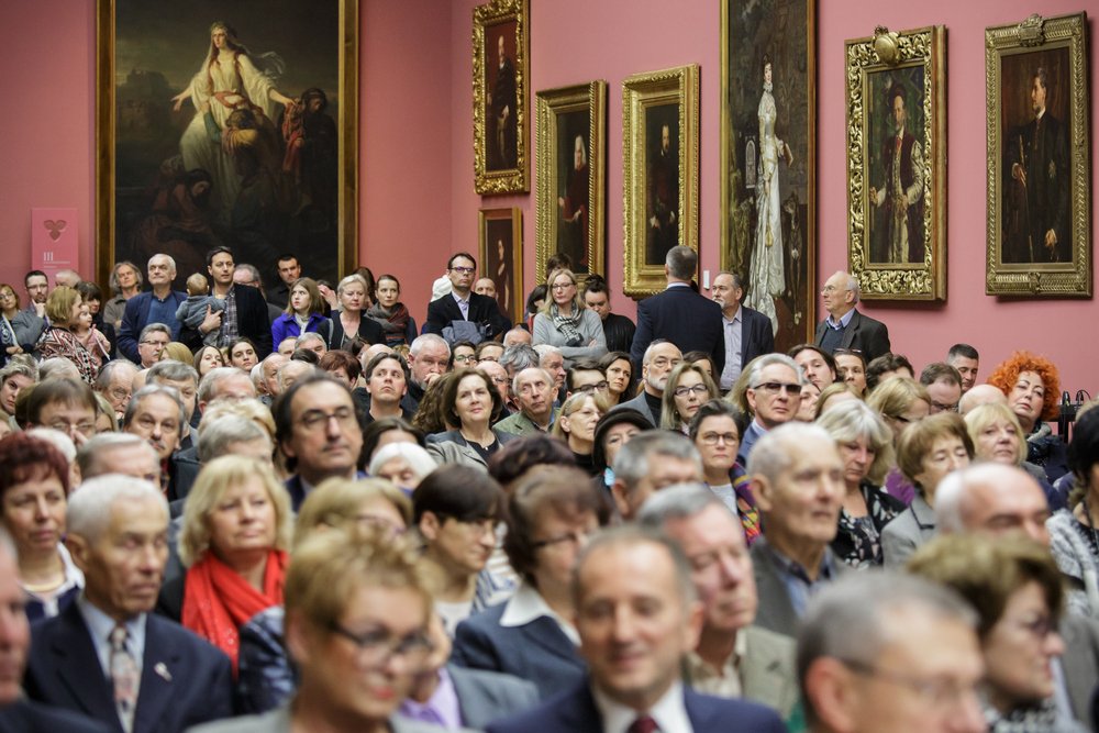 Duża grupa mężczyzn i kobiet w różnym wieku siedzi i stoi w sali wystawowej, a za nimi na różowej ścianie wiszą liczne duże, ozdobne obrazy i portrety w złoconych ramach.