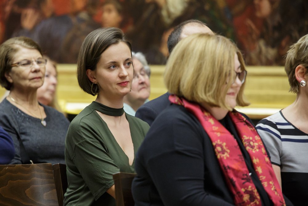 Kilka osób siedzących w rzędach podczas wydarzenia; na środku młoda kobieta o krótkich ciemnych włosach i dużych kolczykach w zielonej bluzce z chokerem, patrząca lekko w bok, po prawej kobieta w okularach i krótkich blond włosach w ciemnym ubraniu z czerwonym, wzorzystym szalikiem, a w tle starsze osoby, drewniane krzesła i duże oprawione malowidło na ścianie.