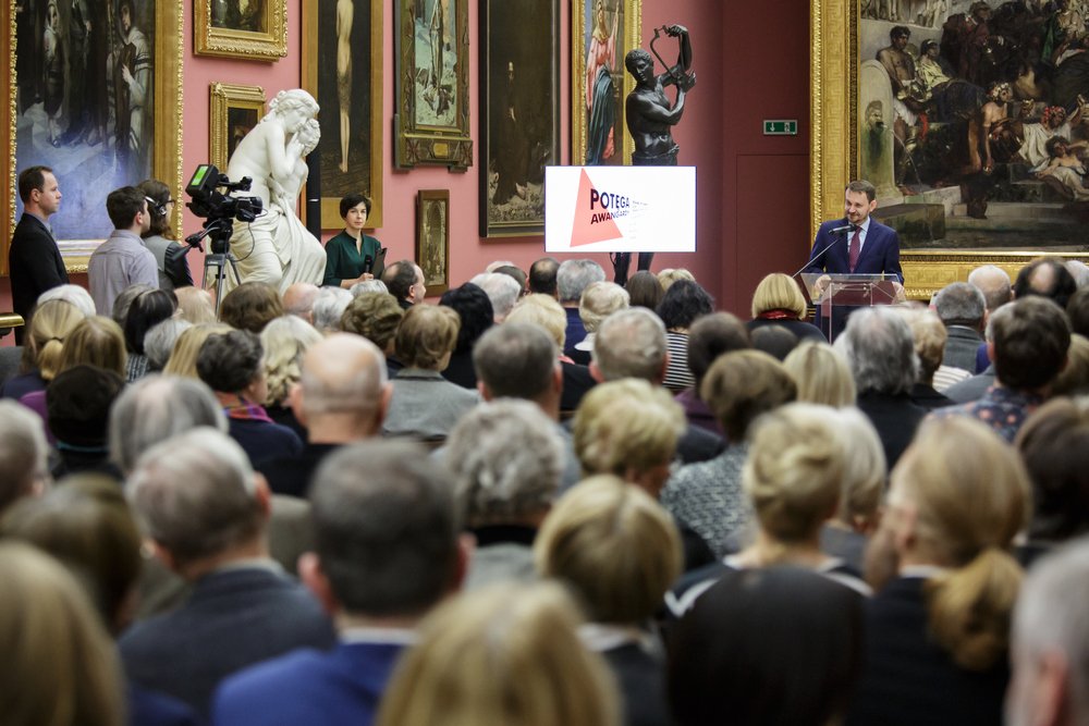 Wnętrze galerii sztuki z dużą widownią siedzącą od tyłu, kamerzystą i ekipą po lewej, białą marmurową rzeźbą i brązową statuą przed ścianą wypełnioną oprawionymi obrazami oraz mężczyzną przemawiającym przy mównicy obok ekranu z widocznym napisem.