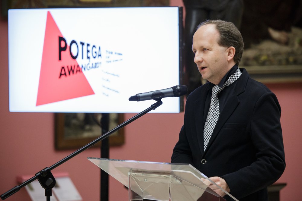 Mężczyzna w ciemnym płaszczu i czarno-białym wzorzystym krawacie stoi przy przezroczystym pulpicie i mówi do mikrofonu na statywie, w tle ekran z czerwonym trójkątem i napisem "POTEGA AWANGARDY" oraz różowe ściany z oprawionymi obrazami.