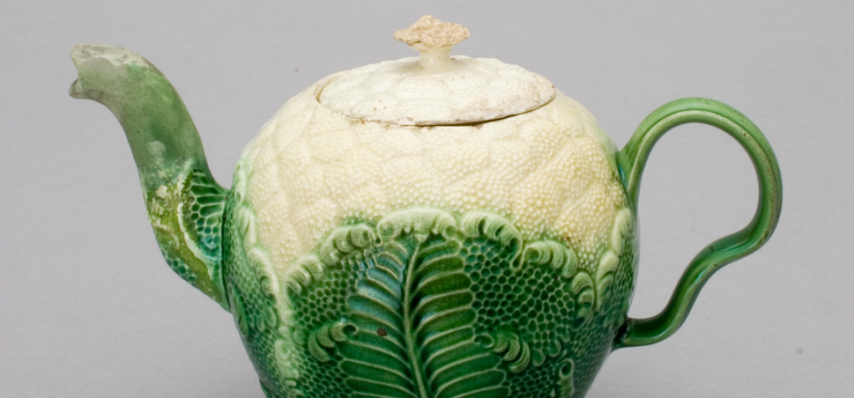 Glazurowany ceramiczny imbryk przypominający główkę kapusty, zielono-kremowy z reliefowym wzorem liści, z pokrywką z małą gałką u góry, wygiętym dzióbkiem po lewej i uchwytem po prawej na jasnoszarym tle.