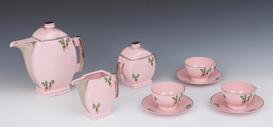 Różowy porcelanowy serwis do herbaty ustawiony na jasnym gradientowym tle: po lewej duży czajnik, obok mlecznik i cukiernica, po prawej trzy filiżanki na spodkach; wszystkie części mają zielone motywy liściowe z małymi pomarańczowymi detalami oraz srebrne metaliczne akcenty przy rantach i uchwytach.