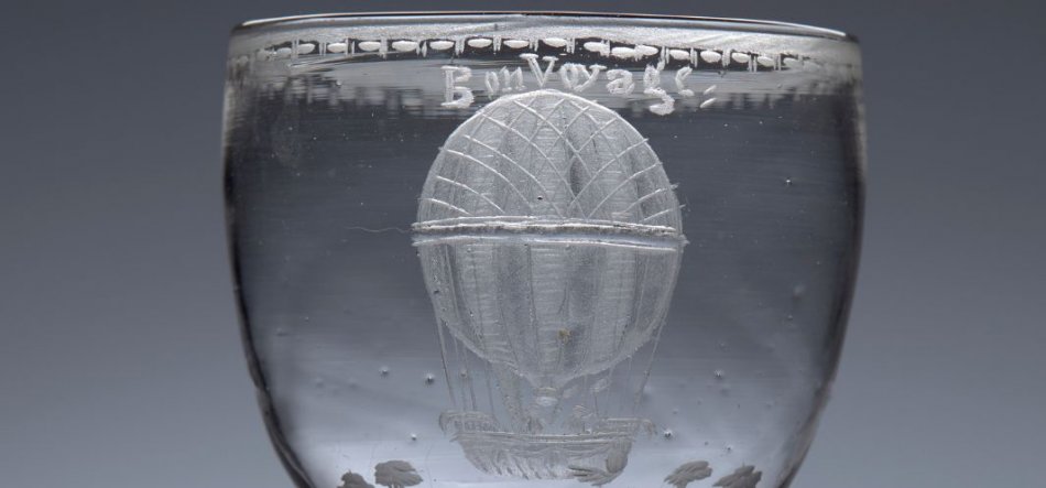 Zbliżenie przezroczystego kielicha ze szkła z centralnym grawerunkiem balonu na ogrzane powietrze z widocznym koszem i linkami, niewielkim napisem nad balonem, pasem wypukłych kropek pod krawędzią i drobnymi grawerunkami roślinnymi przy podstawie.