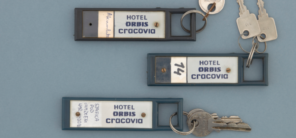 Trzy prostokątne ciemnoniebieskie breloki hotelowe z białymi wkładkami z napisem HOTEL ORBIS cracovia ułożone na jasnoniebieskim tle; dwa breloki mają metalowe kółka z kluczami, jeden ma dopisaną cyfrę 74, a na innym widoczny jest ręczny zapis po lewej stronie etykiety.
