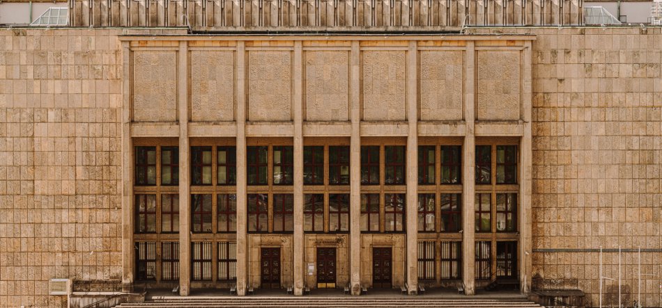 Symetryczna fasada dużego budynku z jasnobeżowych kamiennych płytek, pięć wysokich pionowych przeszkleń rozdzielonych masywnymi filarami, trzy drewniane podwójne drzwi u podstawy oraz szerokie stopnie prowadzące do wejścia, z ozdobnymi panelami nad oknami.