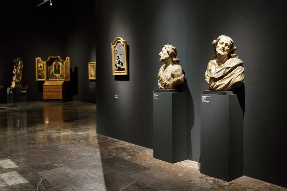 Wnętrze ciemnej galerii muzealnej z czarnymi ścianami i połyskującą marmurową posadzką, na pierwszym planie dwa kamienne popiersia ustawione na wysokich ciemnoszarych piedestałach przy prawej ścianie, w tle po lewej obrazy i rzeźby rozmieszczone wzdłuż ściany.