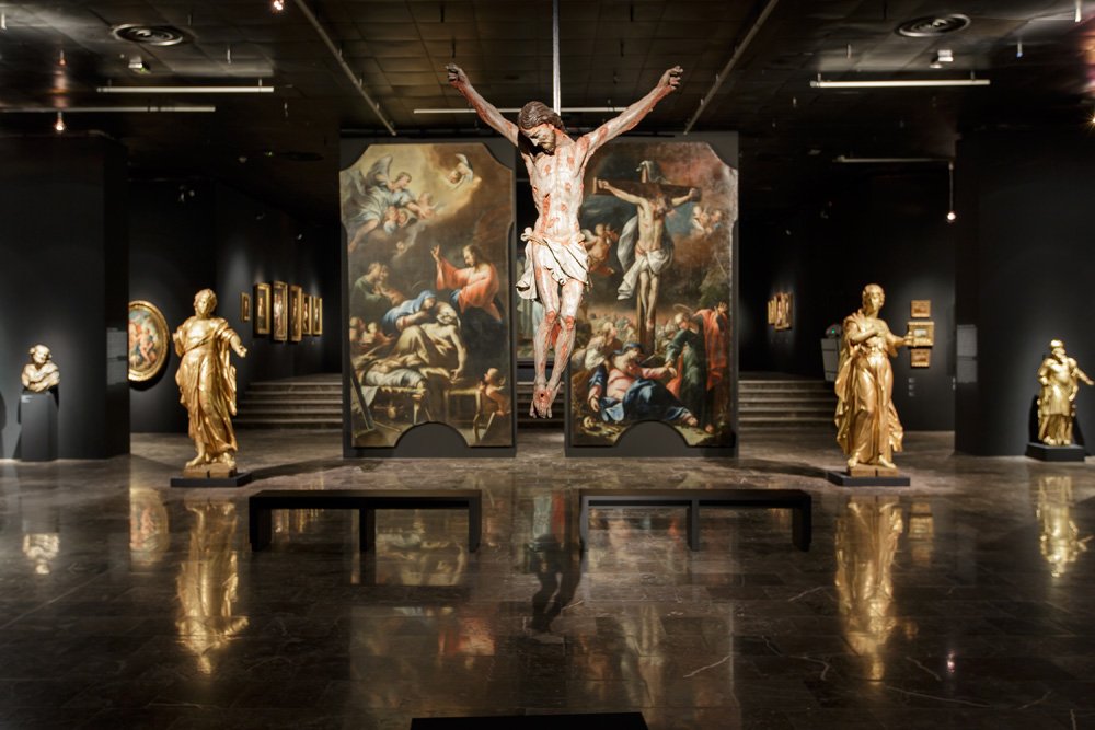 Wnętrze ciemnej sali muzealnej z centralnie zawieszoną rzeźbą ukrzyżowanej postaci przed dwoma dużymi obrazami o tematyce religijnej, po bokach złocone posągi na postumentach oraz dwie ławki na lśniącej, odbijającej podłodze.