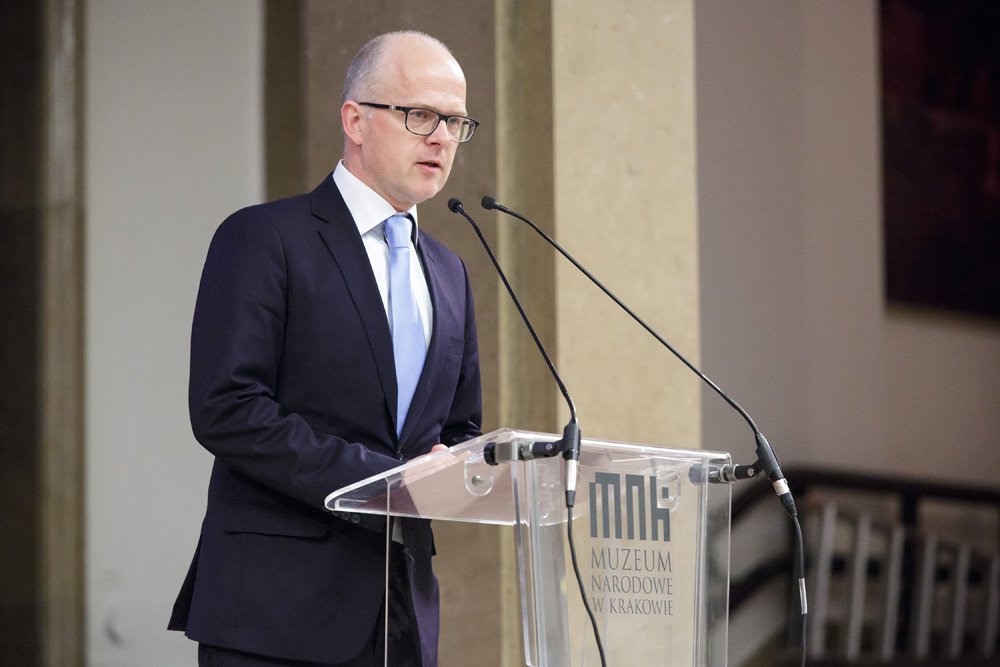 Mężczyzna w ciemnym garniturze i jasnoniebieskim krawacie, w okularach i z krótką fryzurą, stoi przy przezroczystej mównicy z napisem/logo "Muzeum Narodowe w Krakowie" i mówi do dwóch mikrofonów na tle kolumn wnętrza.