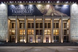 Fasada monumentalnego, wieczorem oświetlonego budynku z wysokimi kolumnami i przeszklonymi drzwiami, nad którymi rozciąga się duży baner z napisem "Skarby baroku" i fragmentem obrazu barokowego przedstawiającym postać.