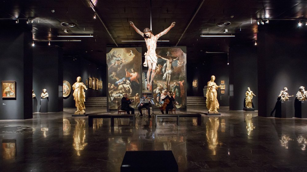 Wnętrze ciemnej galerii sztuki z błyszczącą podłogą, w centrum duża zawieszona figura Ukrzyżowanego na tle wysokich obrazów o tematyce sakralnej, przed nimi siedzący muzycy grający na instrumentach smyczkowych, a po bokach ustawione złocone rzeźby i mniejsze obrazy pod punktowym oświetleniem.