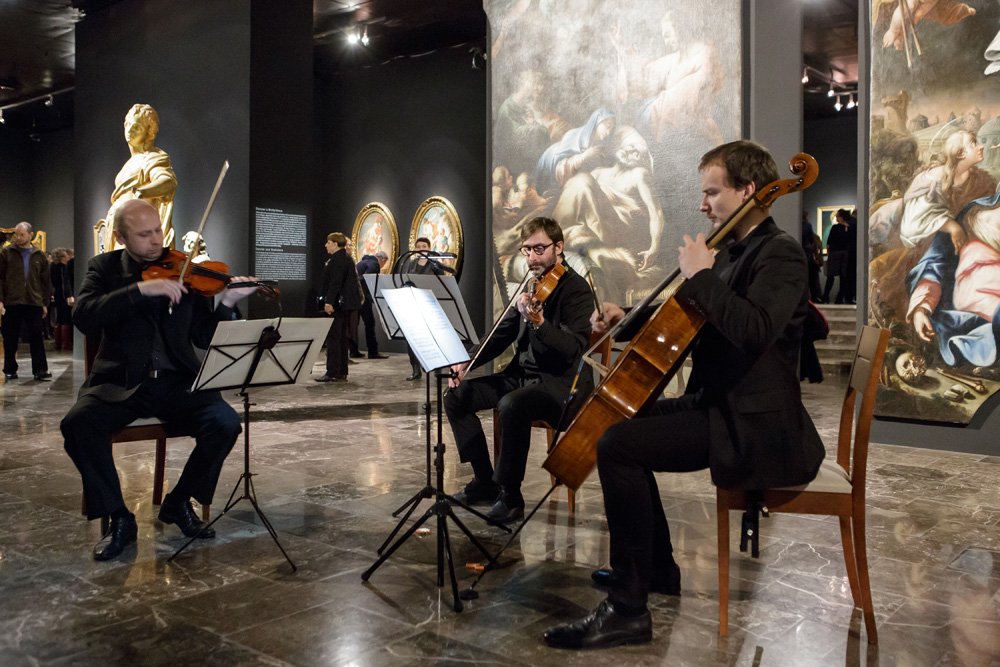 Trio smyczkowe w czarnych strojach siedzi przy pulpitach z nutami i gra na instrumentach na marmurowej posadzce galerii sztuki, wokół duże obrazy i rzeźba oraz zwiedzający w tle.