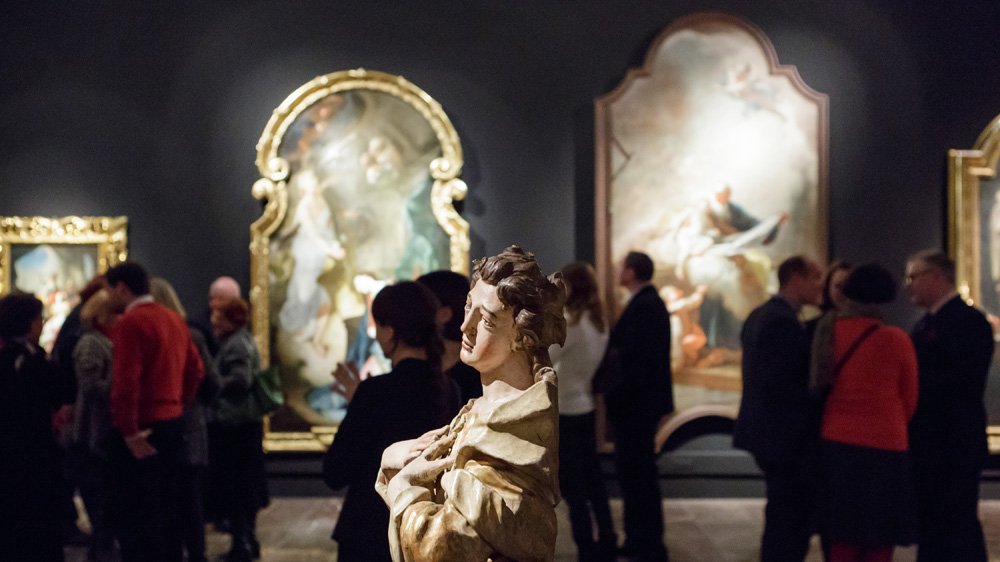 Rzeźba figuralna z jasnego kamienia na pierwszym planie, w tle rozmyte obrazy w ozdobnych złotych ramach i grupa zwiedzających w ciemnej sali galerii.